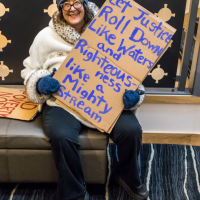 A woman in a blue hat and mittens, and white winter coat, sits on a bench holding a handmade cardboard sign that reads "Let Justice Roll Down Like Waters and Righteousness Like a Mighty Stream".