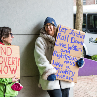 Two women in winter coats lean against against the exterior of a building while holding handmade cardboard signs that read "END POVERTY NOW" and "Let Justice Roll Down Like Waters and Righteousness Like a Mighty Stream".