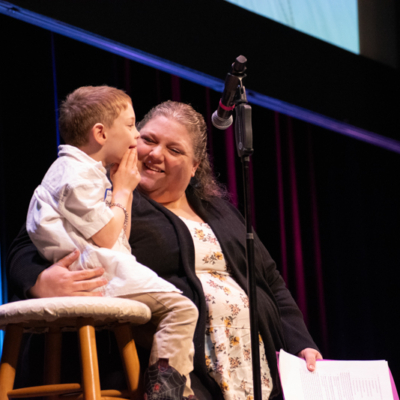 A woman on a stage smiles as she puts her arm around a small boy seated on a stool.