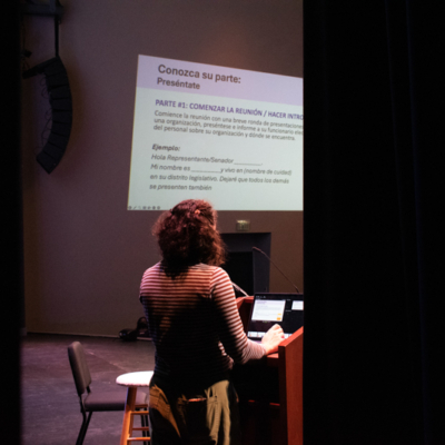A woman speaks at a lectern (photo shot from behind).