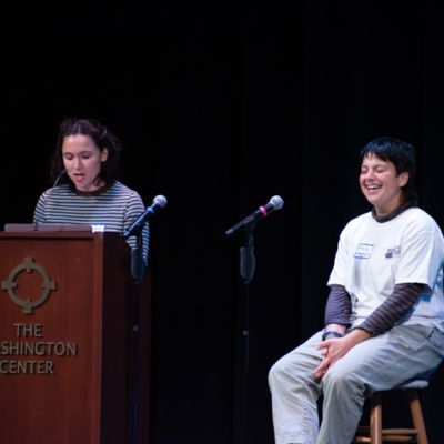 A person seated on a stool laughs as a woman stands at nearby lectern that reads "The Washington Center".