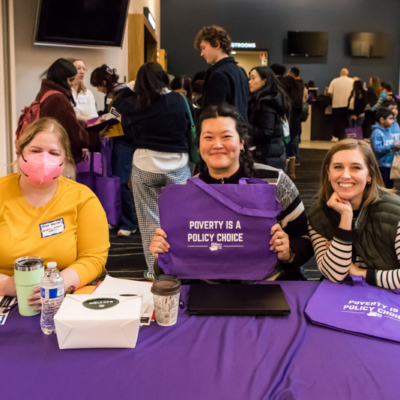 Three smiling people sit at a table in a crowded room, one of them holding up a purple cloth bag that says "POVERTY IS A POLICY CHOICE".