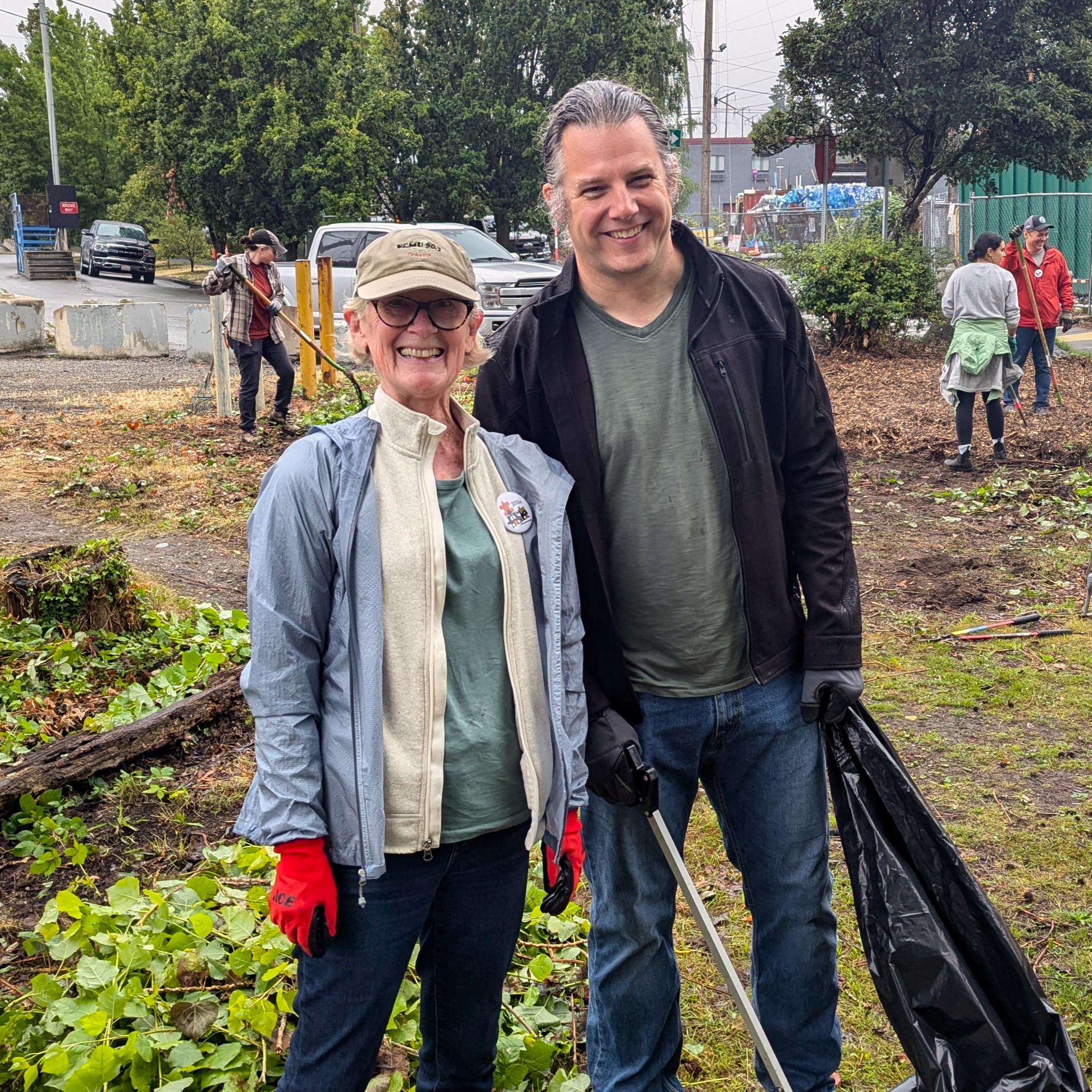 Two people in work clothes pose, smiling, for the camera. One holds a garbage bag and a trash-picker tool. Other people behind them smile as they rake.