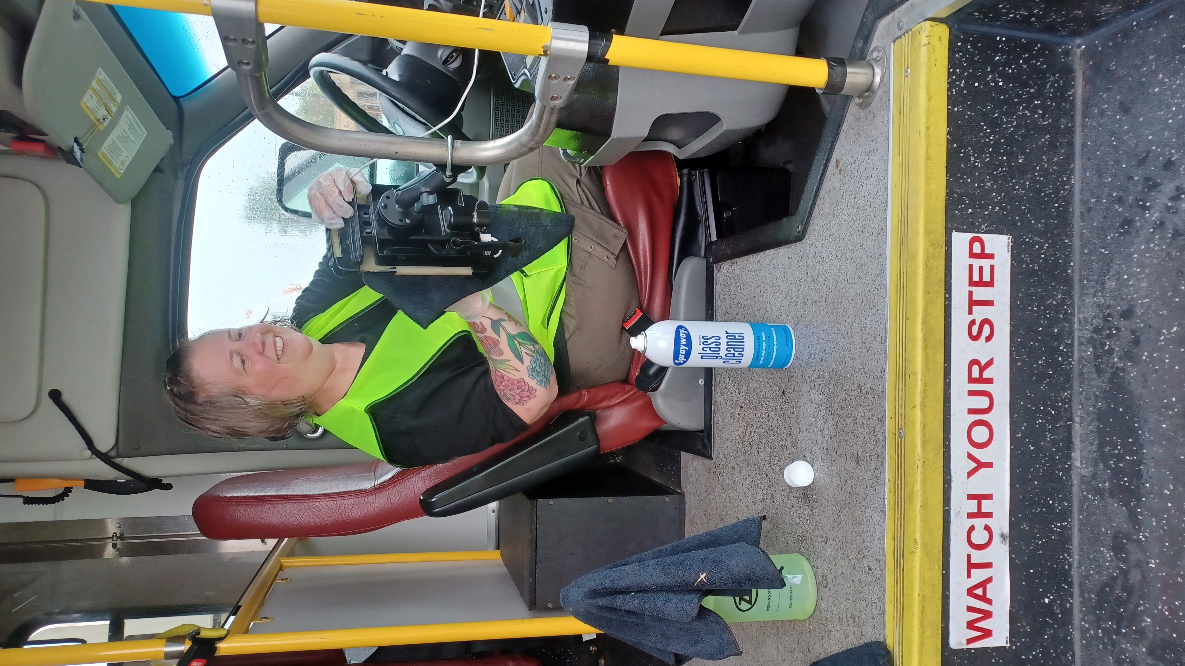 A woman in a neon-yellow vest wipes down the driver's area inside a paratransit bus.