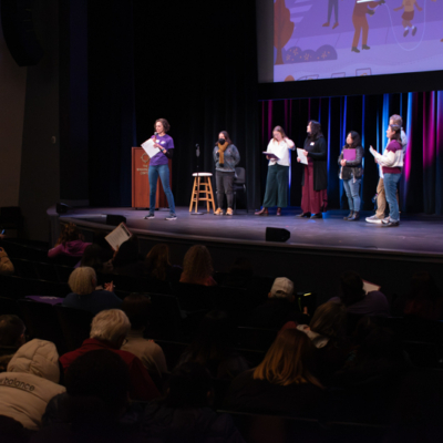 Six people stand on a state, one at a podium, in front of a large audience in a dark room.