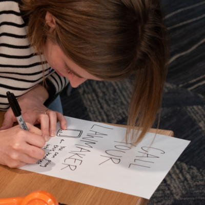 A woman leans over a sheet of paper with a black permanent marker and writes on a hand-drawn sign that starts with "Call your lawmaker" followed by smaller text.