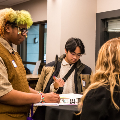 A man with a coat slung over his shoulders clicks a pen as two others face him.