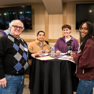 Four people stand around a circular high-top table covered in papers and water bottles.