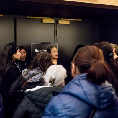 A group people listen to a woman speaking in front of a set of black doors.