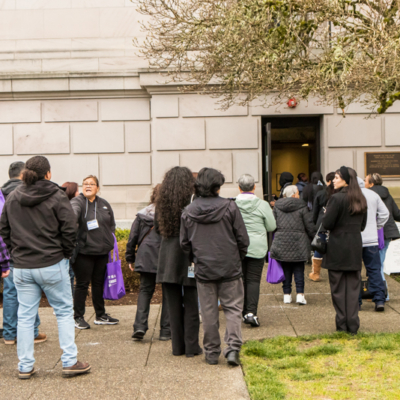 A large group a people stand on a sidewalk as some of them file into a door next to a bronze plaque on a building.