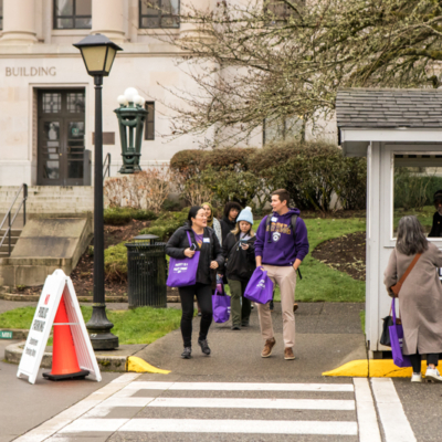 Several people with purple bags step into a crosswalk next to a small security kiosk.