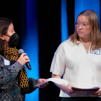 A woman in a black mask speaks into a handheld microphone while onother woman looks at her.