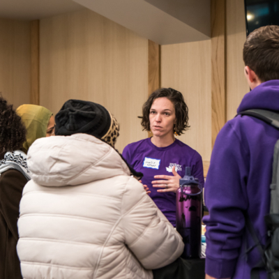 A woman in a purple shirt gestures as she talk to two people in winter coats with their backs to the camera.