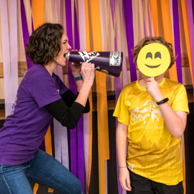 A woman in a purple shirt pretends to scream through a blowhorn directly at a small boy in a yellow T-shirt who's covering his face with a yellow smiley face prop.