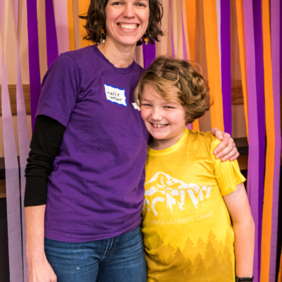 A woman in a purple shirt hugs a child in a yellow T-shirt with one arm while holding a purple blow horn in the other.