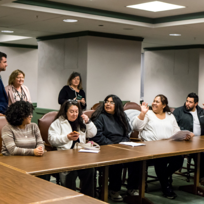 Several people seated along a wooden table raise their hands while looking up at a man standing off to the side.