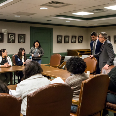 Two men in suits stand at the end of U-shaped table while several people sit in chairs along its edges.