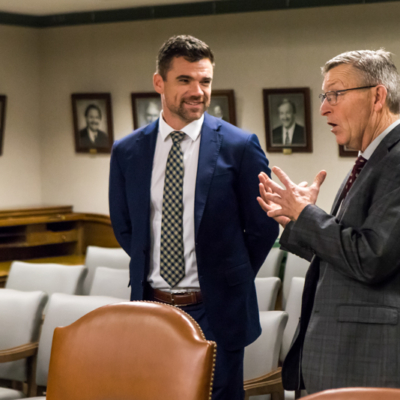 A man in a suit speaks as he gestures with his hands while another man in a suit and tie looks on.