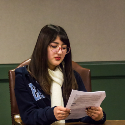 A woman in a scarf sits at the edge of a wooden table and carefully studies a sheet of paper in her hands.