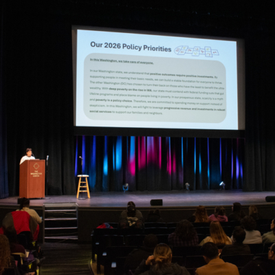 A speaker stands at a podium on a stage next to a large screen that reads "Our 2026 Policy Priorities" followed by a long paragraph of text.