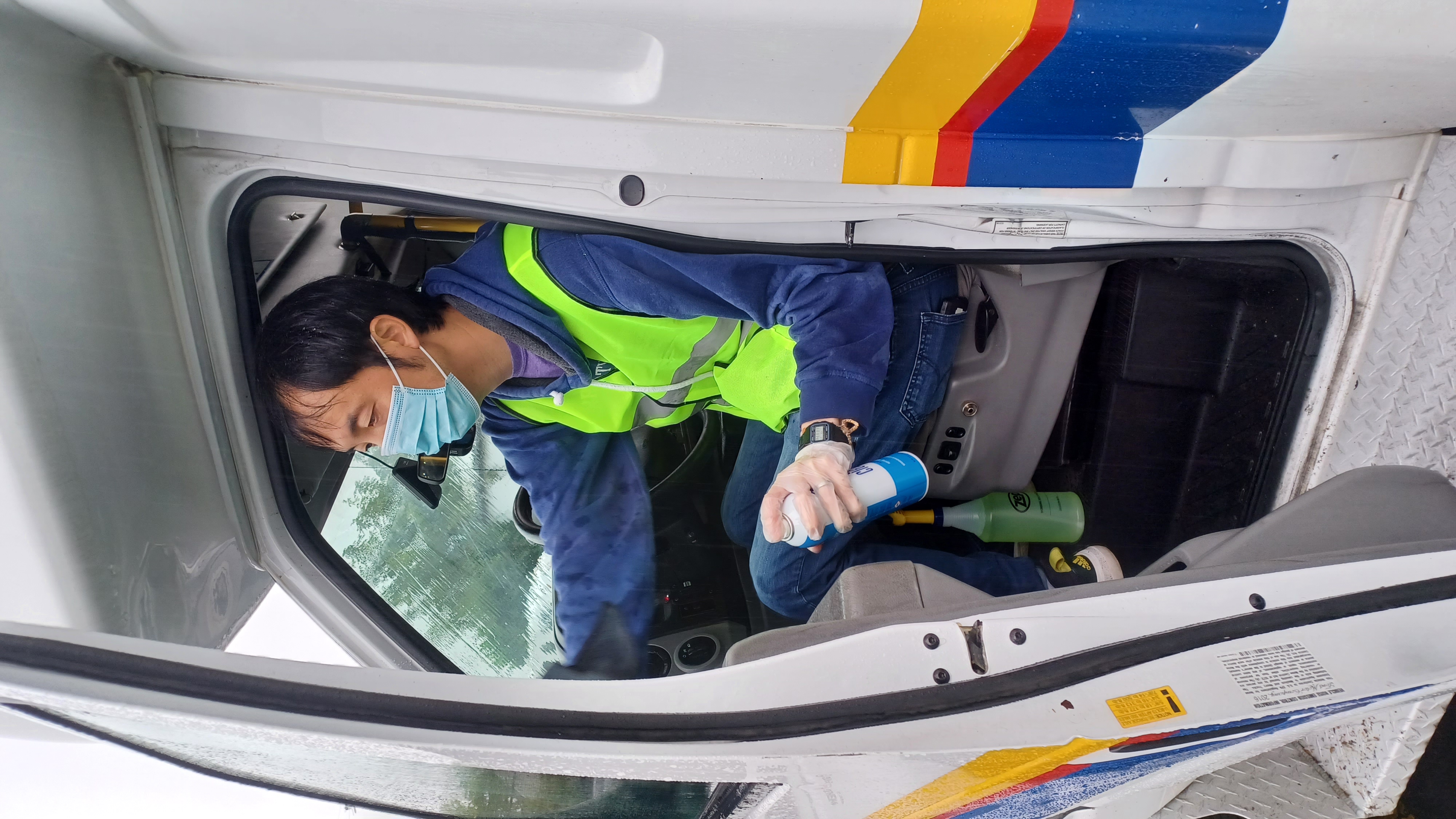 A man in a yellow neon vest and wearing a blue face mask cleans windows inside a large van.