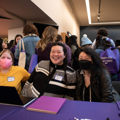 Three people sit at a long table in front of a group of other people.