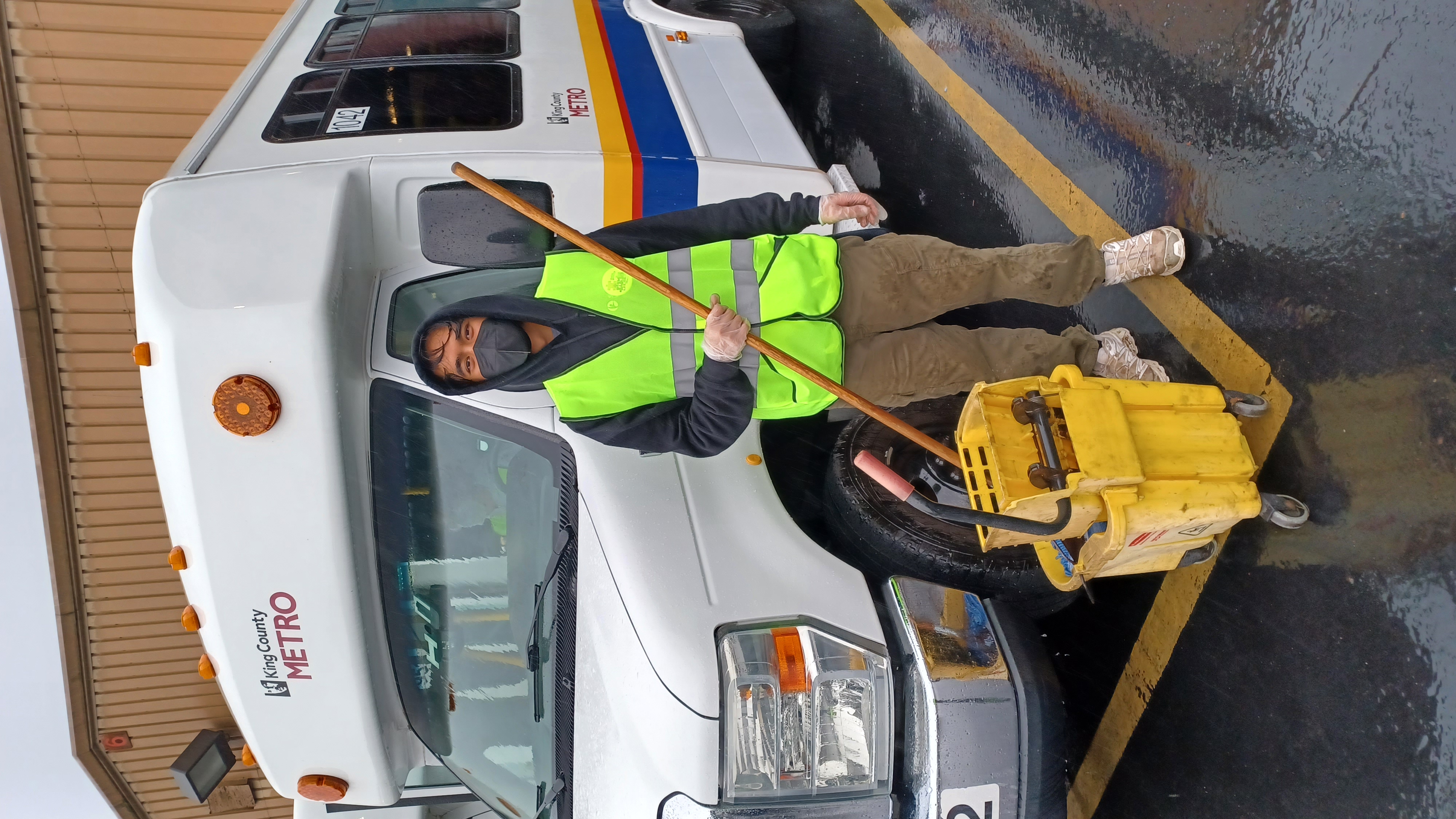 A man in a neon-yellow vest stands with a mop and bucket in front of a paratransit van.