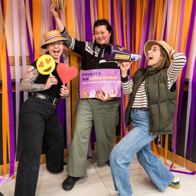 Three people strike goofy poses while holding props, including a sign that says "Poverty is a policy choice."