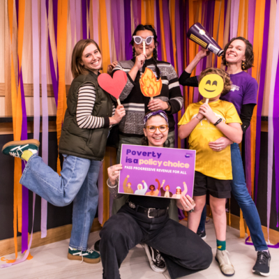 Five people strike goofy poses while holding props, including a sign that says "Poverty is a policy choice."
