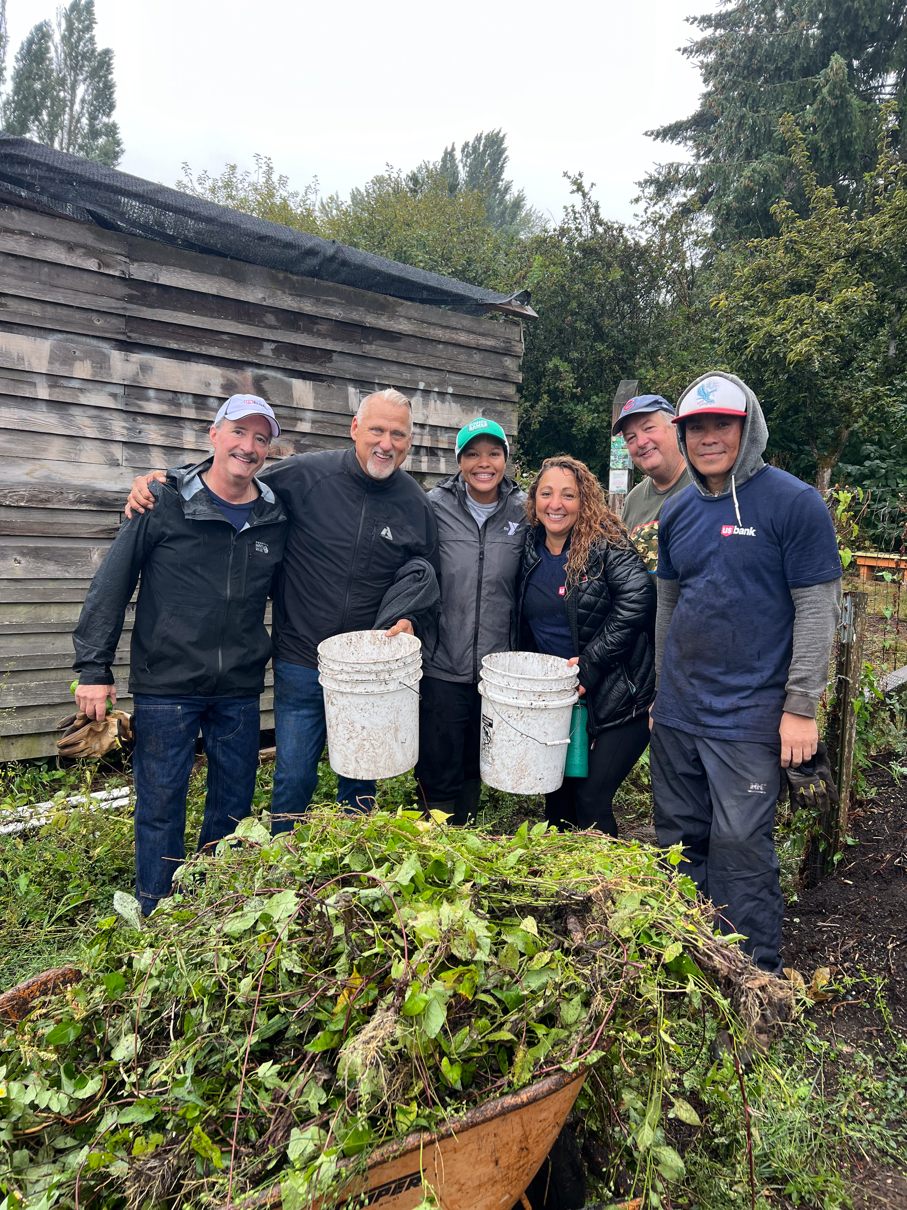 A group of people, some of them holding white five-gallon buckets, stand over a large pile of weeds.
