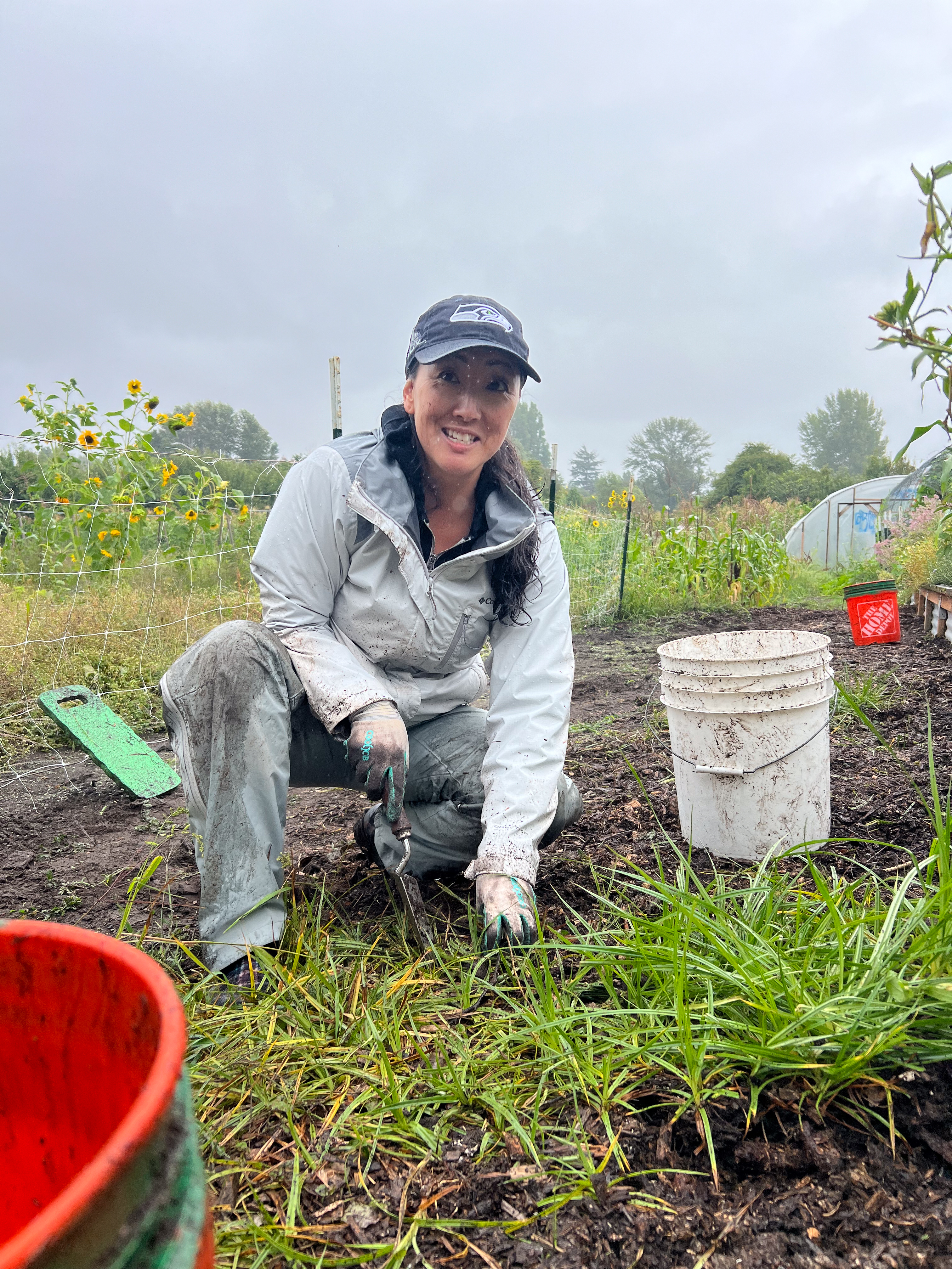 A woman in a blue rain jacket and jeans smiles while crouched down pulling weeds