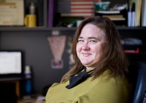 A woman with dark brown hair and an olive-green sweater leans back in her desk chair and smiles at the camera. In the background and out of focus are two computer and a variety of items on a shelf.