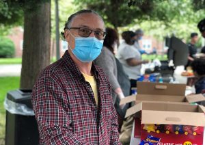 A man wearing surgical mask, sunglasses, and a plaid shirt stands in a row of people handing out food at a BBQ.