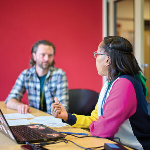A man with facial hair and a flannel shirt consults with a woman in glasses and a multicolor top. They sit at a conference table with a laptop and paperwork. The wall behind them is red.