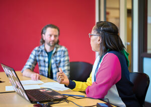 A man with facial hair and a flannel shirt consults with a woman in glasses and a multicolor top. They sit at a conference table with a laptop and paperwork. The wall behind them is red.