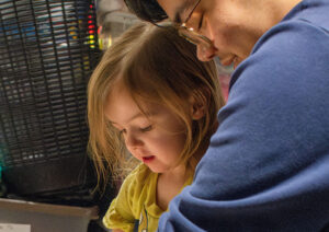 A tight shot of a young girl with wispy blond hair with an adult in a blue sweater behind her helping her with something out of frame.