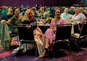 A group of people in fancy dress sitting around a table in a large, crowded ballroom, several them turned in their seats to face the camera.