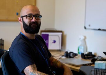 A man in a full, dark beard and blue tshirt looks at the camera and smiles while seated an office desk.