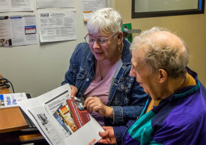 Two gray-haired adults talking to each other while holding and pointing to Medicare brochures.