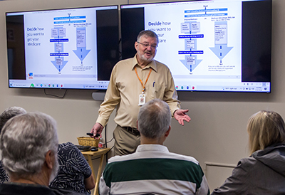 A man stands in front of a pair of screen showing information about Medicaid while several people look on.