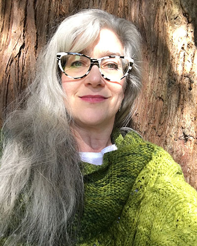 Selfie portrait of a woman in front of a large tree trunk. She has long white hair, glasses, and wears a green sweater.