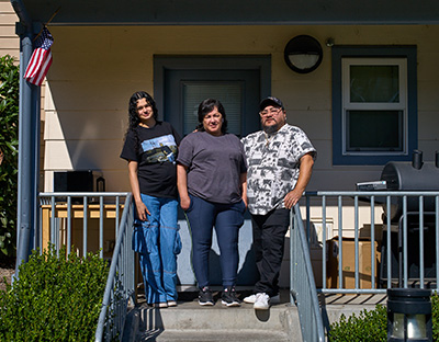 A couple and their daughter stand at the top of the stairs to a small porch infont of a home.