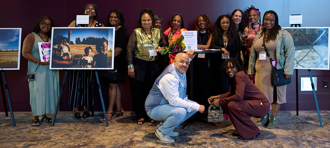 A large group of family and friends of a photographer pose smiling with joy, surrounded by large photo posterboards of her work.