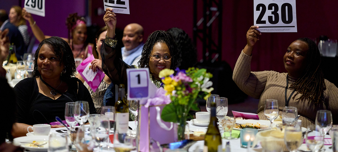 A group of smiling people in fancy dress raise bidder numbers high at a fancy dinner table.