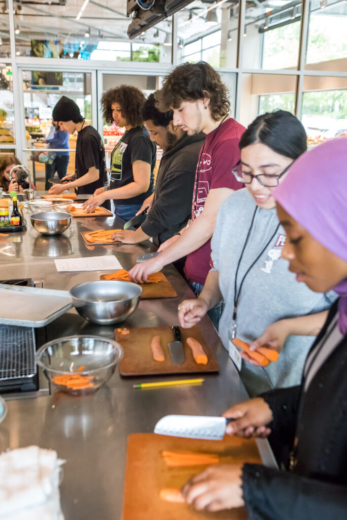 A row of cooking class students practice julienning carrots on individual wooden cutting boards.