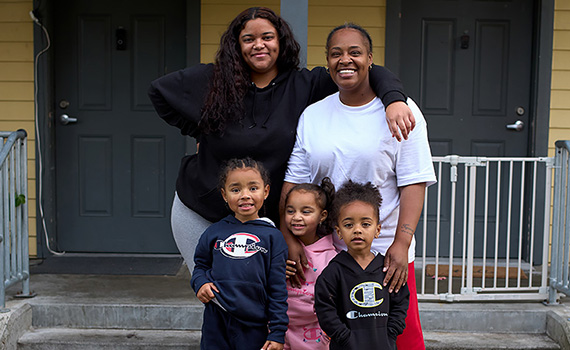 Two moms stand in front of their shared front porch with their 3 kids in front of them.
