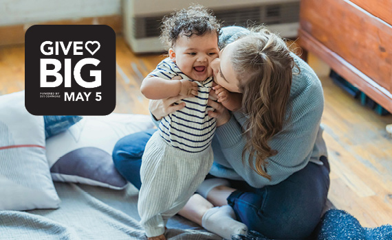 Mom kisses the cheek of her grinning, curly-haired baby as they sit on the floor together. To their left is a black and white graphic reading GIVE♥BIG MAY 5.