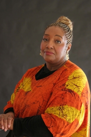 Portrait of Bettie J. Williams-Watson, a Black woman with braids swept up in a bun, wearing an orange, yellow, and black tunic