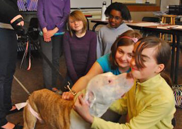 Five middle schoolers smile as one girl in a yellow shirt gets "kisses" from a dog.