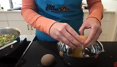 Video screenshot of a woman's arms cracking an egg into a measuring cup